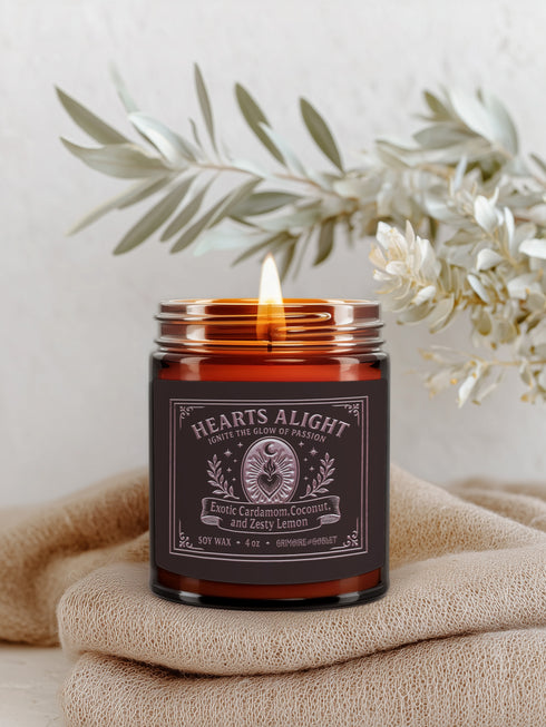 A circular, dark amber glass jar candle with a solid gold-colored metallic lid. The glass jar has a large, rectangular black label with a light-colored decorative border and ornate scrollwork. The label features the title "Hearts Alight" at the top in elegant script and lists the scent notes "Exotic Cardamom, Coconut, and Zesty Lemon" below it. A single white cotton wick with a lit flame is visible, centered within the wax. The wax itself is a rich, warm, orange-amber color, consistent with the jar's hue.